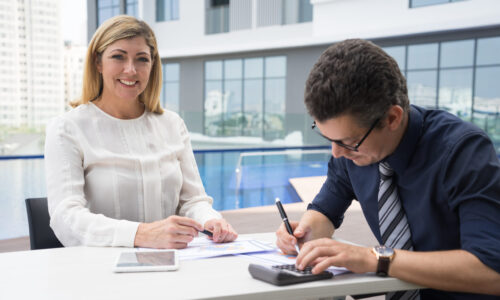 Cheerful excited mature female financier smiling at camera while accountant using calculator in outdoor business cafe. Successful business executive meeting with financial advisor. Informal meeting concept